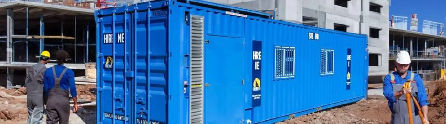 Oficina de contenedor de embarque azul instalada en un sitio de construcción con empleados cerca.