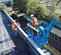 Vista frontal en ángulo de dos empleados con equipos de seguridad operando un brazo de elevación Genie azul en un estacionamiento en la azotea.