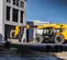 Vista lateral de un manipulador telescópico JCB amarillo con un aditamento para montacargas moviendo material de construcción en un sitio de trabajo exterior.