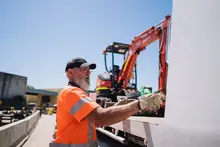 Worker standing beside a loader truck at a construction site, inspecting or adjusting equipment near safety barriers.