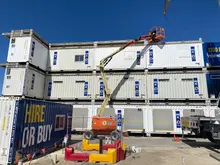 Construction site with shipping containers used as offices and storage, surrounded by safety barriers and workers in high-visibility gear.